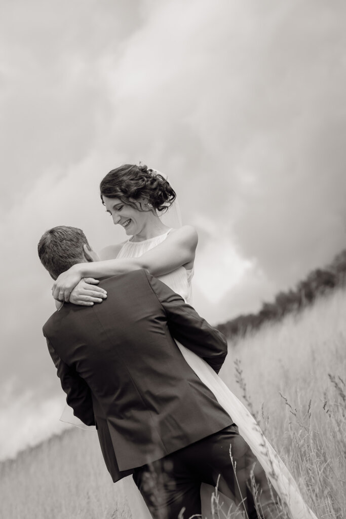 Hochzeitsfoto des Brautpaares auf einem Feld, wo der Brautigam die Braut hoch hebt in schwarz weiß