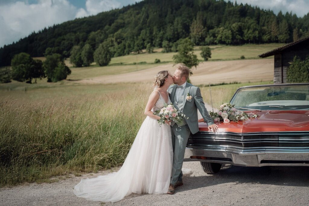 Brautpaar steht auf einem Feldweg mit Auto und küsst sich