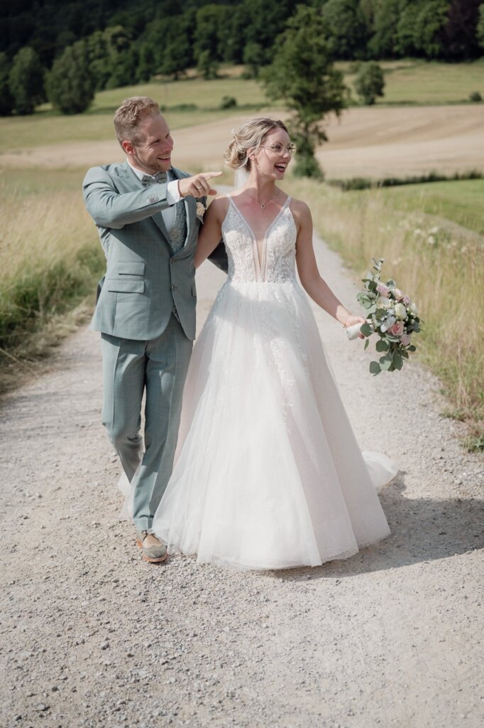 Das Brautpaar geht einen Feldweg entlang und er zeigt ihr etwas in der Ferne, worüber sich beide freuen