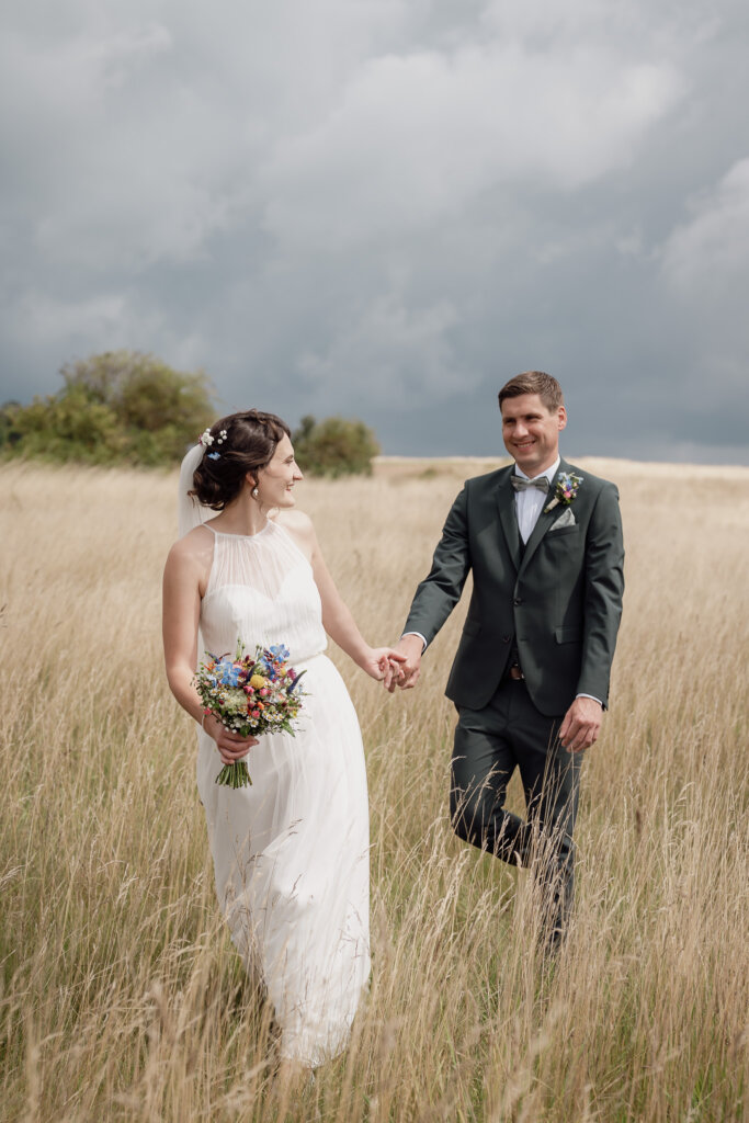 Brautpaar geht über das Feld schaut sich dabei an und die Braut zieht Braeutigam hinterher