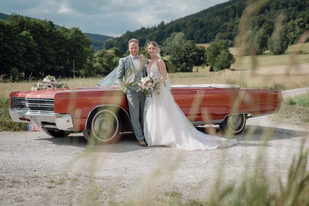 Hochzeitsfoto von dem Brautpaar an einem Oldtimer mit Blick in die Ferne