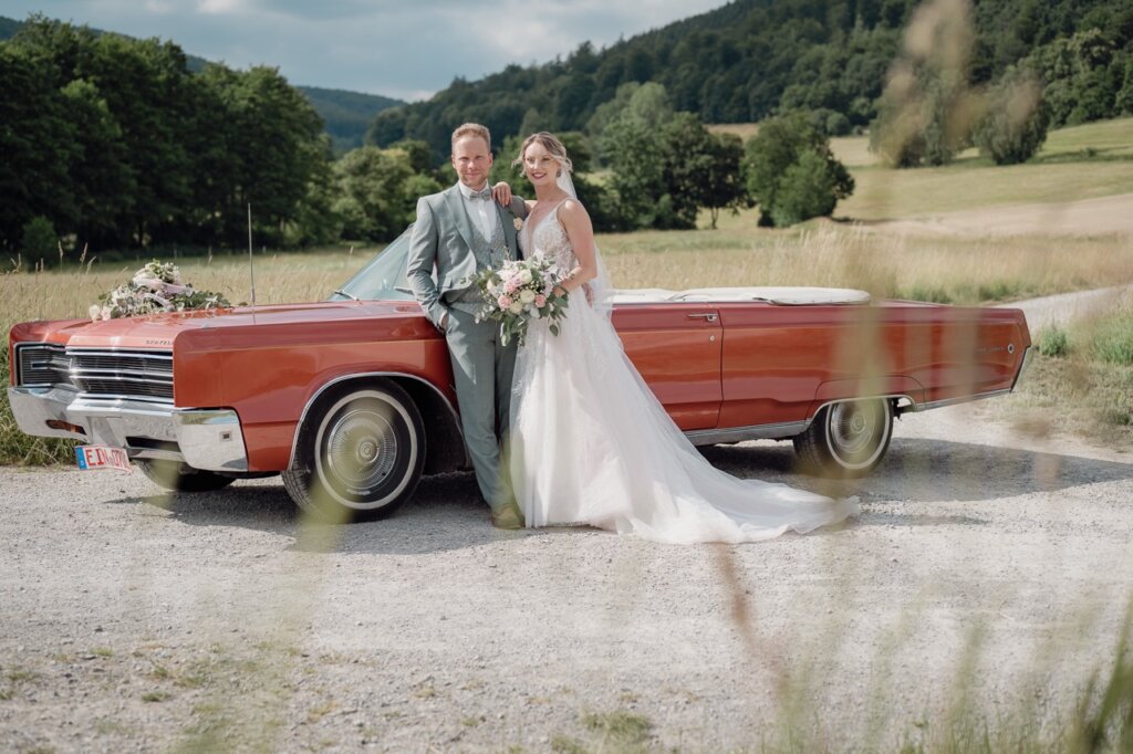 Hochzeitsfoto von dem Brautpaar an einem Oldtimer mit Blick in die Ferne
