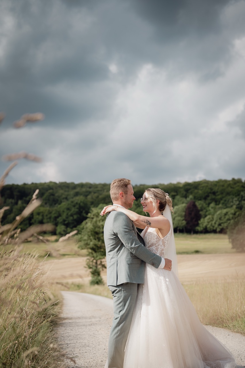 das Brautpaar steht im Sonnenschein auf einem Feldweg und sie legt ihre Arme um seinen Hals während im Hintergrund düstere Wolken aufziehen