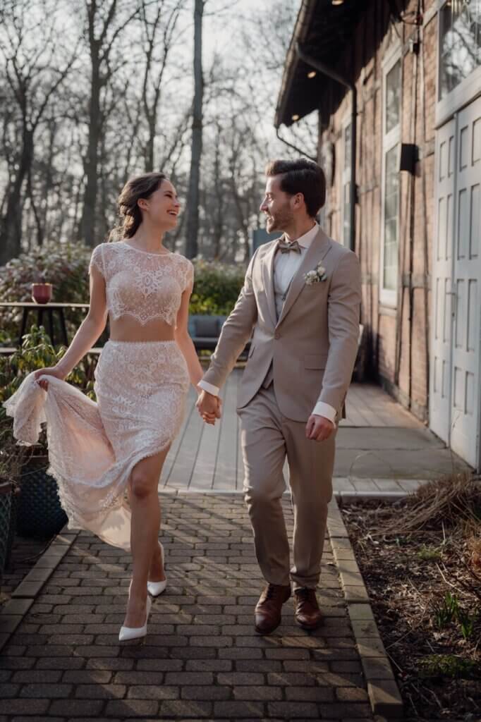 Brautpaar geht gemeinsam an dem Peak Hotel entlang und lächelt sich an, während sie ihr Kleid hoch hält